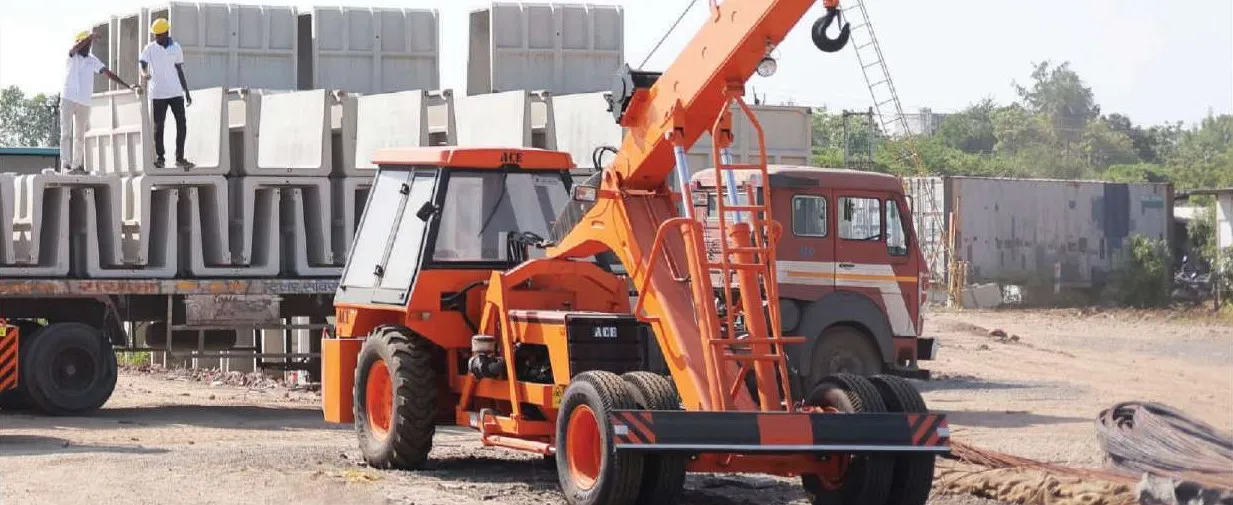 An orange ACE crane beside a truck trailer stacked with precast U-shape drain units, two workers in hard hats standing on top of the load