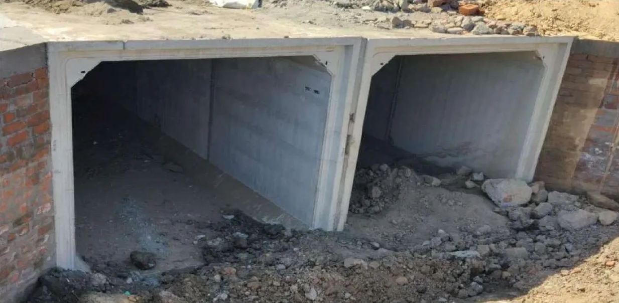 An installed twin-cell precast box culvert set between brick walls, its openings partly filled with soil and rubble