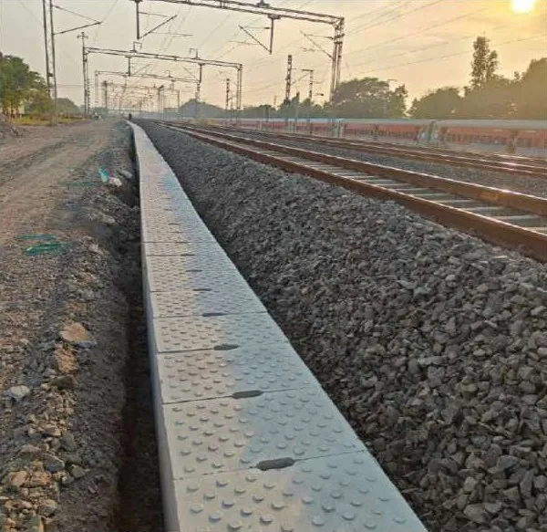 A line of precast drains closed with studded cover slabs running beside a ballasted railway track under overhead electric lines