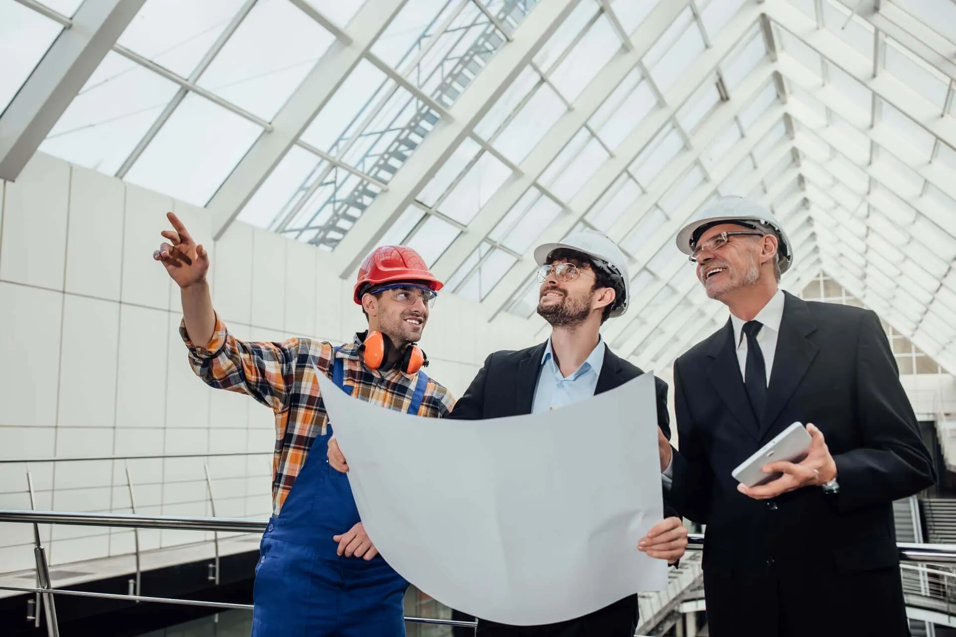Three engineers in hard hats reviewing a large drawing under a glass roof, one pointing upward