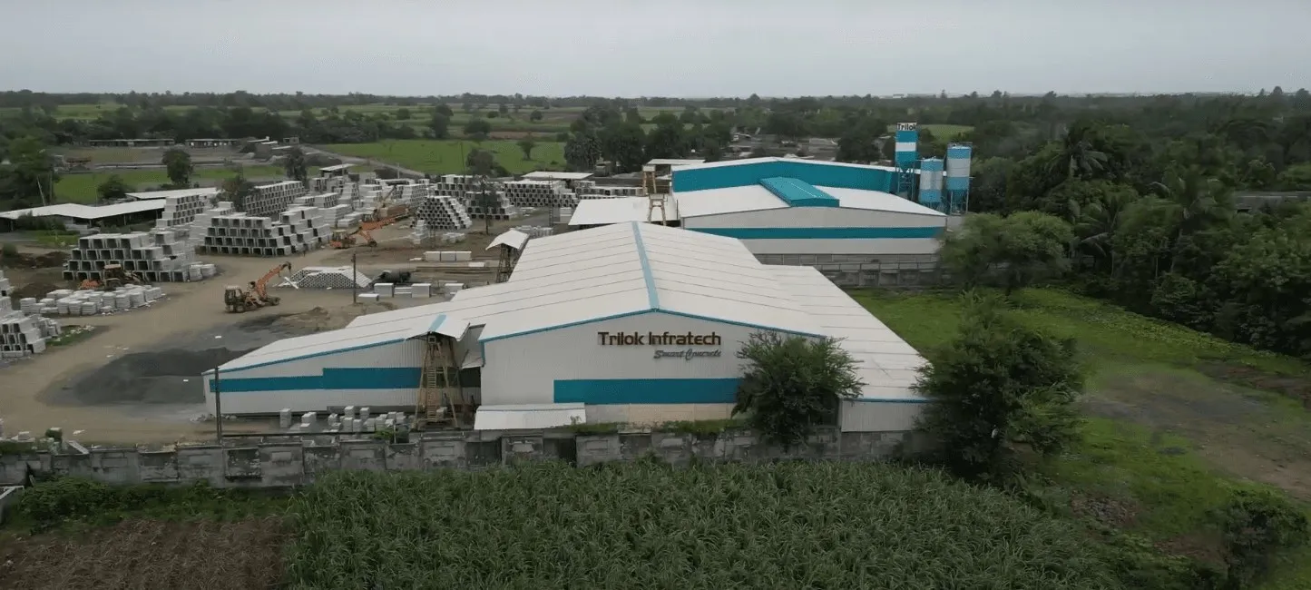 Wide aerial view of the Trilok Infratech plant among farmland: the signed main shed, a second teal-roofed shed with cement silos, and a large yard of stacked precast drains and culverts