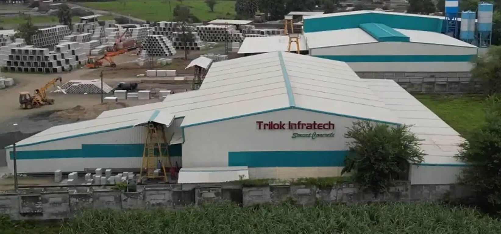 Aerial view of the Trilok Infratech precast plant: a white-and-teal factory shed signed “Trilok Infratech – Smart Concrete”, with a yard of stacked precast units, a crane, loaders and cement silos behind it