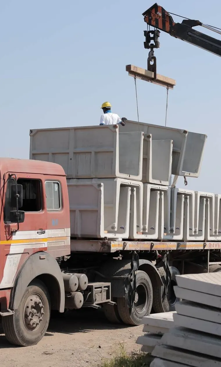 An orange mobile crane loads precast U-shape drain units onto a flatbed truck while a worker in a hard hat guides them; stacks of drain cover slabs lie in the foreground