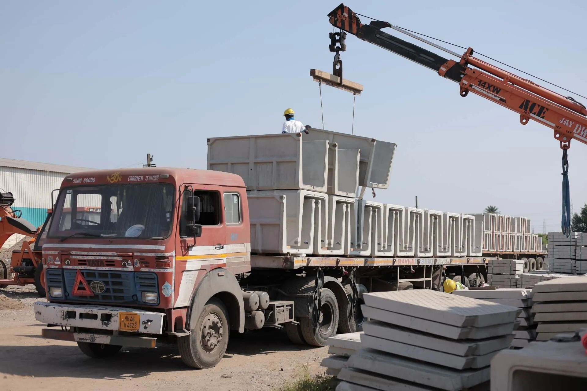 An orange mobile crane loads precast U-shape drain units onto a flatbed truck while a worker in a hard hat guides them; stacks of drain cover slabs lie in the foreground